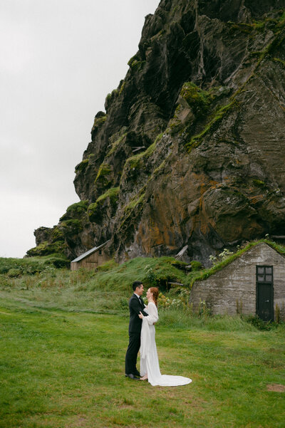 With joy, the bride lifts a champagne glass near a table adorned with a tower of sparkling glasses by the house