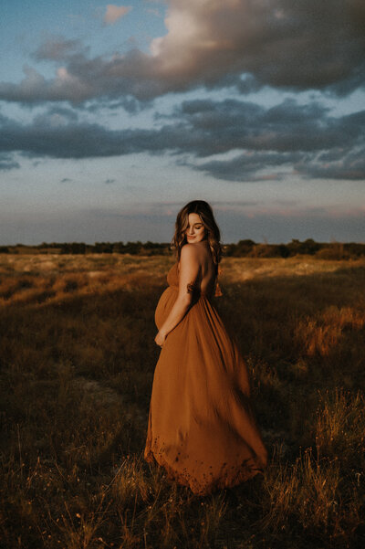 A pregnant mom standing in a field in decatur, tx holding her belly with blue clouds in the background