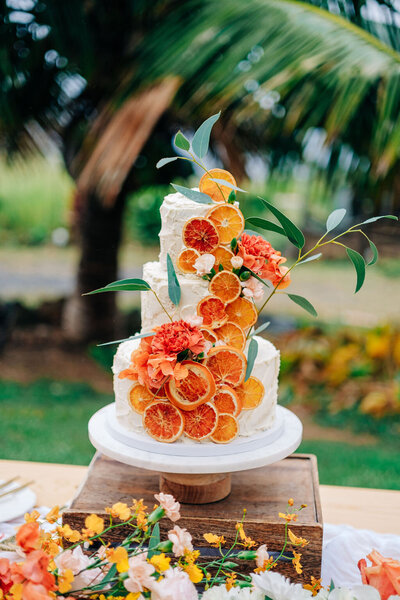 Elegant wedding cake adorned with dried orange slices, set on a floral-filled table featuring tropical and traditional blooms. Created by Chef Jasmine of HeartBeet Catering at Puuwai Gardens and beautifully captured by photographer James Rubio