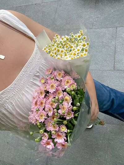 A person in a white dress holds bouquets of pink and white daisies, standing on a gray tiled pavement. The scene conveys a fresh and cheerful mood.