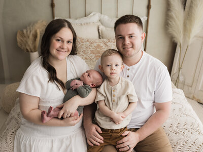 mom and dad sitting on bed holding 2 year old son and newborn baby boy for studio portraits