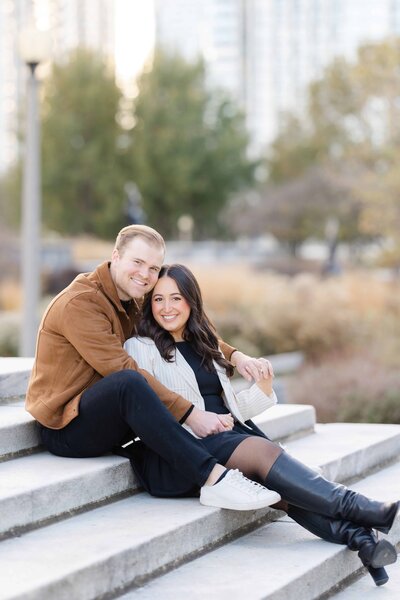 Man in brown leather coat sitting with woman in between his legs.  The woman is wearing a white jacket with knee high black leather boots at the Chicago Museum Campus in Autumn for their photos by Winterlyn Stebner, an engagement photographer in Chicago