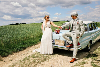 Ein hübsches Brautpaar in cremefarbigem Hochzeitsgewand sitzt auf dem hellblauen Mercedes Oldtimer. Beide tragen Sonnenbrillen und lachen sich an.