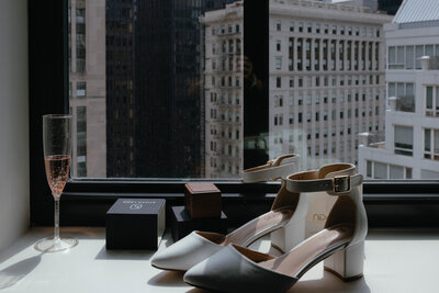 A pair of white kitten heels with ankle straps sits next to a glass of champagne and ring boxes near a window overlooking skyscrapers in Chicago.