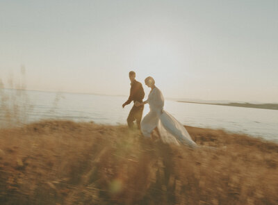 bride and groom running through a field by the ocean