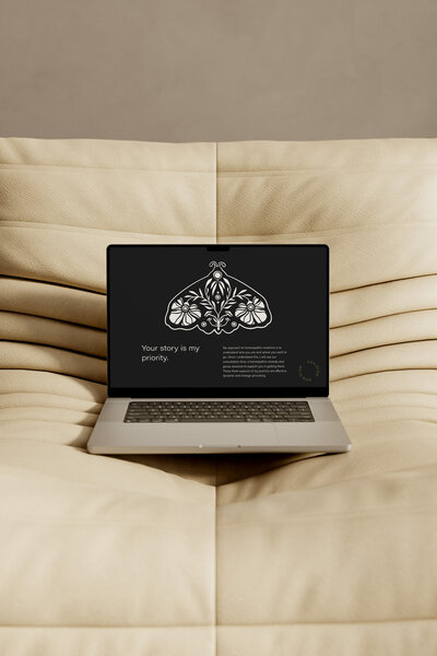Laptop on a beige sofa displaying a black screen with a floral moth illustration and the text 'Your story is my priority.'