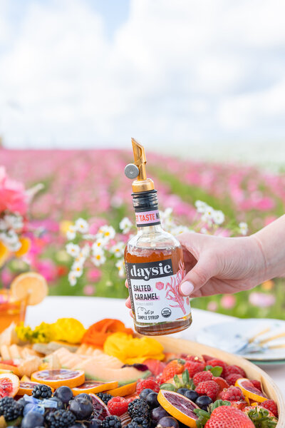 Daysie syrup being held over a plate full of delicious, vibrant fruits in a flower field. I traveled from Charleston, SC to photograph this brand shoot with a glassware company.