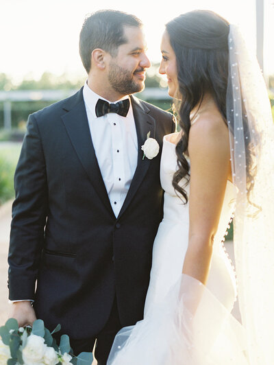 Bride and groom smiling at each other while groom holds bride's bouquet