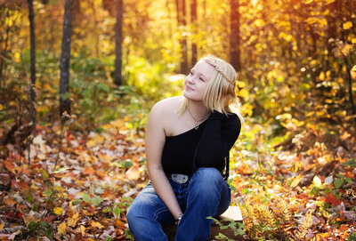 high school senior girl sitting on the ground looking off smiling with a vibrant yellow glow from the fall colors behind her in Akeley Minnesota