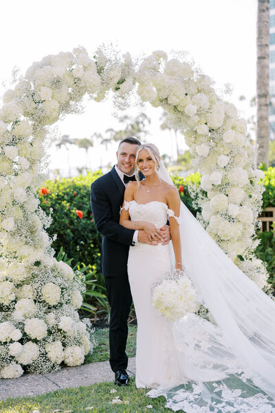 Bride and groom embracing and smiling at the camera with floral arch in the background