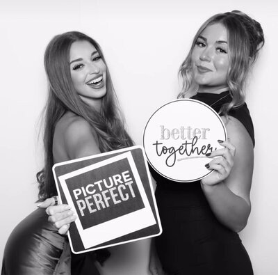 Two women smiling and posing in front of a white backdrop. One holds a square sign that reads “Picture Perfect,” while the other holds a round sign that says “Better Together.”