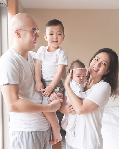 asian family in white tee shirts and light grey sweats in their beige master bedroom holding 3 year old son and new baby girl