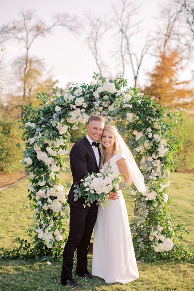 Bride and groom standing under lush floral arch with ivory and greenery accents