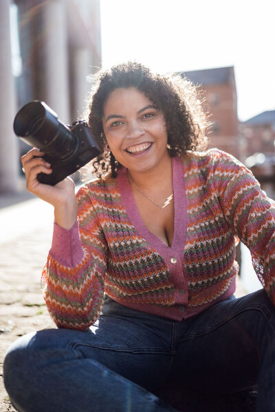 Mirlah  the photographer holds a camera wearing a pink striped cardigan