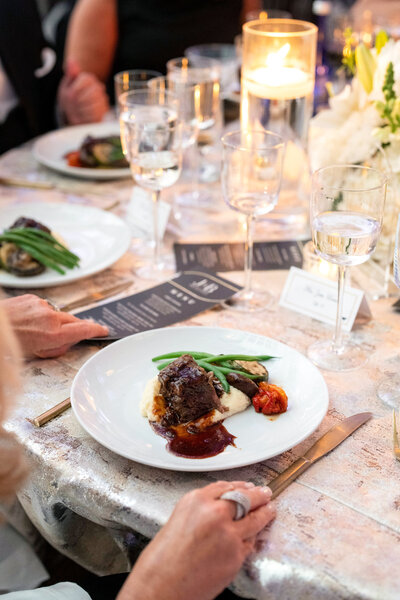 A plated gourmet meal on a table with a lit candle and wine glasses. The dish includes a piece of meat with sauce, green beans, mashed potatoes, and a roasted cherry tomato. Elegant place setting with a printed menu and name card nearby.