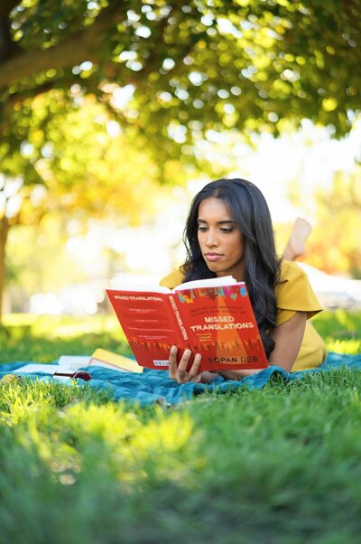 Tania, wearing a yellow dress, reading a red book on a blue blanket in the middle of a natural setting with trees in the background