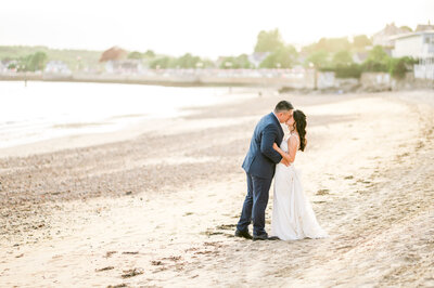 A bride and groom share a kiss on the sandy beach beside the Beauport Hotel in Gloucester, MA, with soft golden light and gentle waves creating a romantic coastal scene.