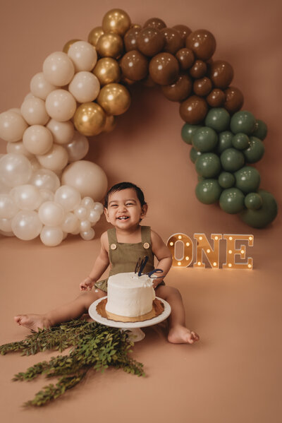 One year old brown haired baby boy sitting on tan backdrop and wearing a olive green romper eating a smash cake with Marietta GA balloon garland decoration at an Atlanta portrait studio