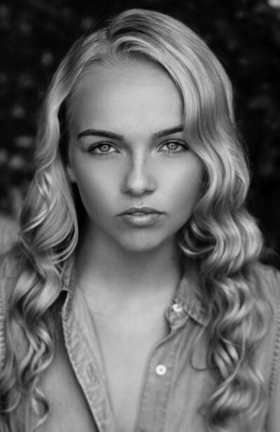 Black and white portrait of a young woman with long wavy blonde hair, wearing a button-up shirt, and looking directly at the camera with a calm expression.