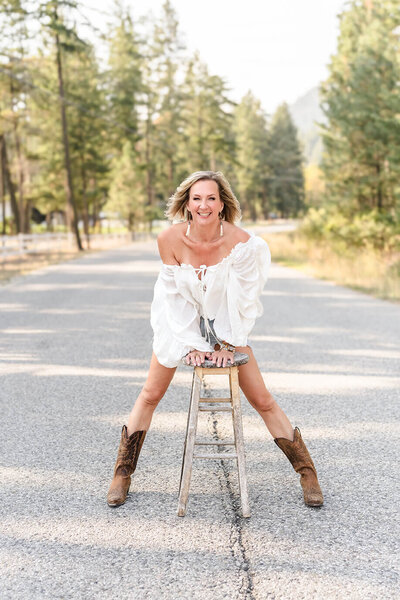 Pam Rader wearing white top and shorts and leaning forward on stool in the middle of a country road.
