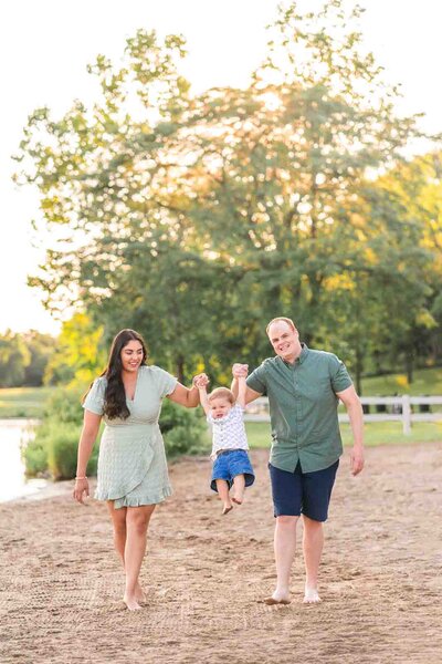 Mother and father swinging two year old boy back and forth on the beach with sunset in background