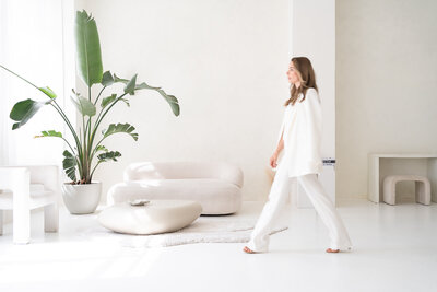 žena v bílém kostýmku přechází v pohybu přes světlou místnost, v pozadí je pohovka, velká rostlina v květináči a konferenční stolek