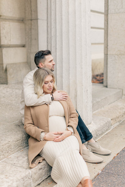 Expecting couple sitting on stone steps wrapped in a warm embrace during a maternity session in Kansas City.