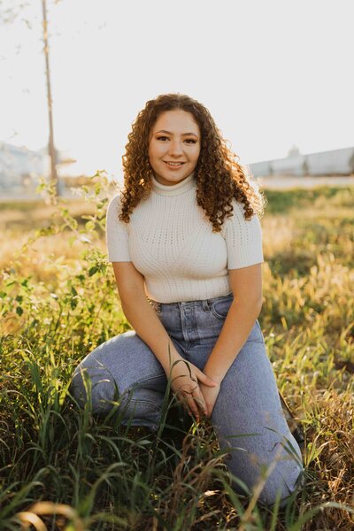 curly haired senior girl sitting in downtown West Bottoms in Kansas City at sunset. Smiling at camera with golden light hitting her hair and making the background glow.