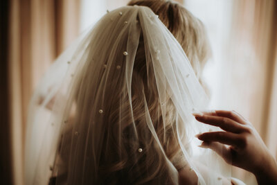 Bride holding a pearled veil while looking out the window.