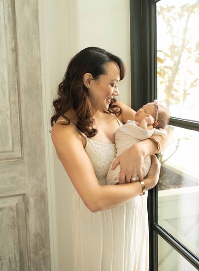Mother in an elegant dress cradles her newborn near a bright window. A serene newborn session by Moments in Light Photography.