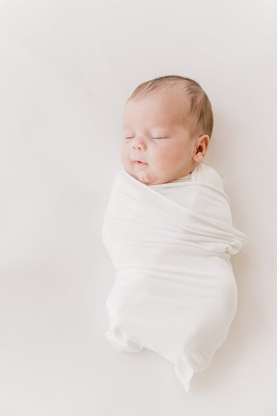 A newborn wrapped in a white swaddle on a white background.