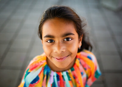 Portrait of a smiling young girl with dark hair, wearing a vibrant, multicoloured top, captured in natural light on a paved outdoor surface in Kingston, Ontario.