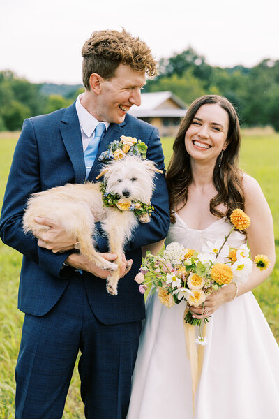 A bride holding a bouquet smiles next to the groom holding their dog wearing a floral collar in the mountains of North Carolina, by film photographer Megan Lynn of My Sun and Stars Co.