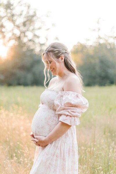A pregnant woman wearing a pink dress stands in a field holding her belly.
