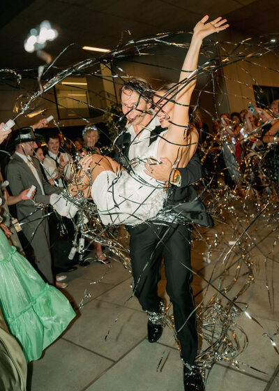 Flash photo of groom carrying bride through the line of their wedding guests while silver streamers fly around them
