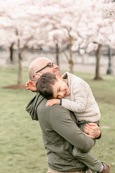 Dad holding young son with cherry blossom trees in the background. They are both smiling very big. Dad is wearing an olive green shirt with a hood and the boy is wearing green pants and a cream sweater. Portland Spring Cherry Tree Family Photos.