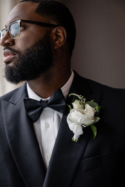 a black groom in a black tux with a bowtie and boutonniere staring out the window