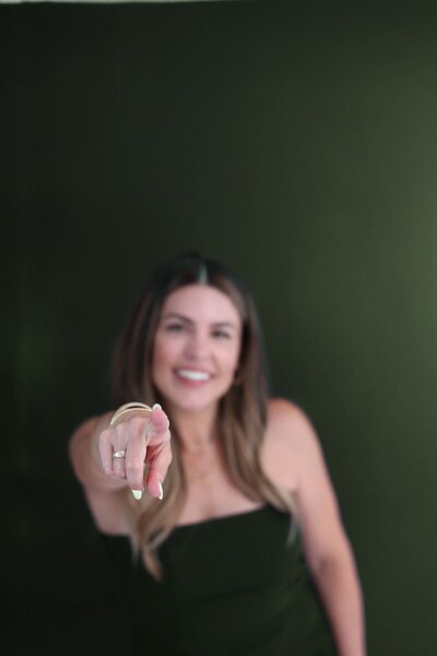 Smiling woman in black outfit pointing directly at the camera against a dark green background, encouraging personal empowerment.
