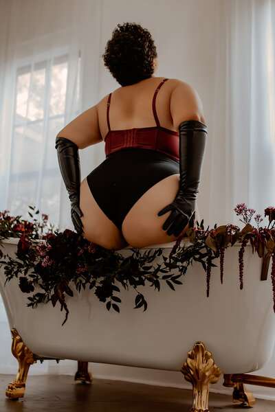 Boudoir photography in Cleveland, Texas featuring a woman in a red corset sitting on a floral-decorated clawfoot bathtub
