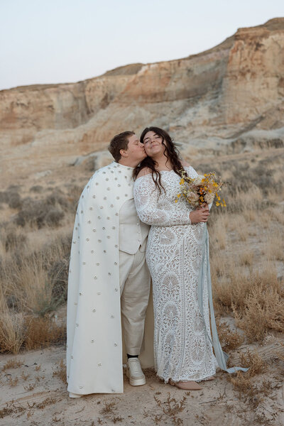 Two brides embrace at their LGBTQ Luxury wedding at Amangiri Resort in Utah. One bride is wearing a white bedazzled suit cape. The other bride is wearing a Grace Loves Lace Gown and is holding an organic yellow bridal bouquet.