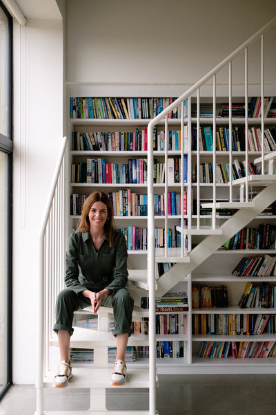 A woman sits on a white staircase, surrounded by tall bookshelves filled with books.