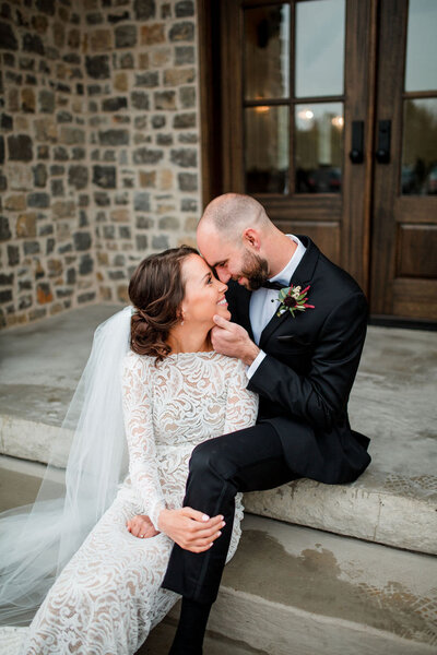 Groom lovingly holding brides chin so she can look up at him while they are snuggled together on stairs outside of Birchwood