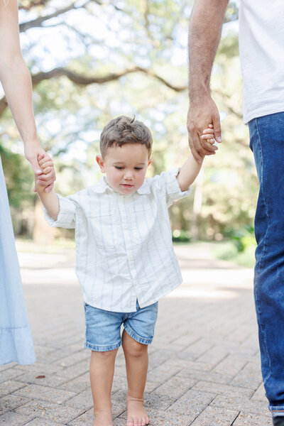 A toddler boy holding his mom and dad's hands while walking at Eden Gardens State Park during a family photography session..