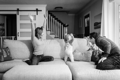 A black and white image of a family at home, where the parents and their baby are playing on a couch. The parents have their arms raised, mirroring their baby’s joyful gesture.