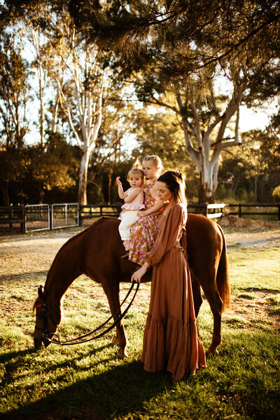 A woman stands beside a brown horse while holding the reins, with two young children sitting on the horse's back, surrounded by trees in a grassy area.