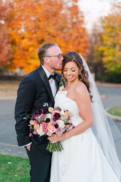 A groom kisses his bride on her temple with colorful orange trees behind them.