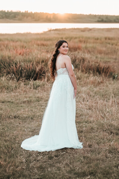 BRIDE LOOKING OVER HER SHOULDER AND SMILING WHILE STANDING IN A GRASSY FIELD WITH THE SUNSET BACKGROUND in san antonio, Texas.