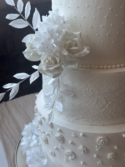 Cloe-up photo of all white wedding cake with white roses, stencil tier background, and pearls
