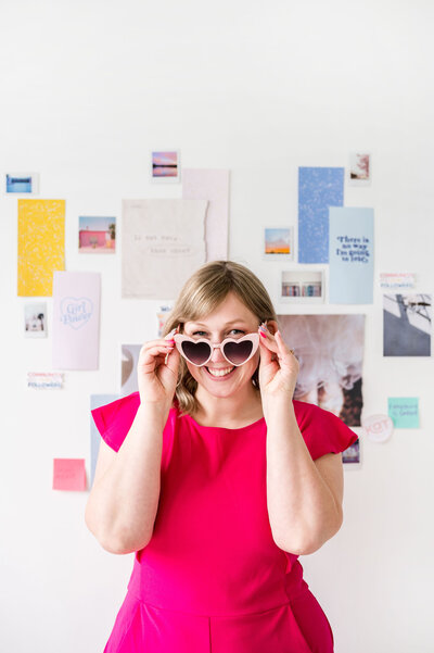 Kat Murphy in a pink jumpsuit putting on pink heart sunglasses in front of a collage wall