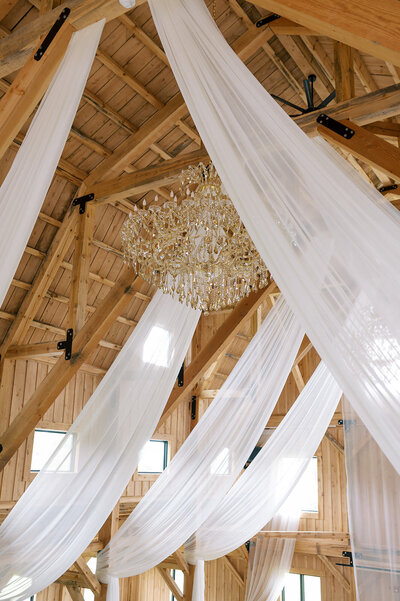 Elegant barn ceiling draped with flowing white fabric and a crystal chandelier, creating a romantic ambiance at Bear Ridge Destination.

